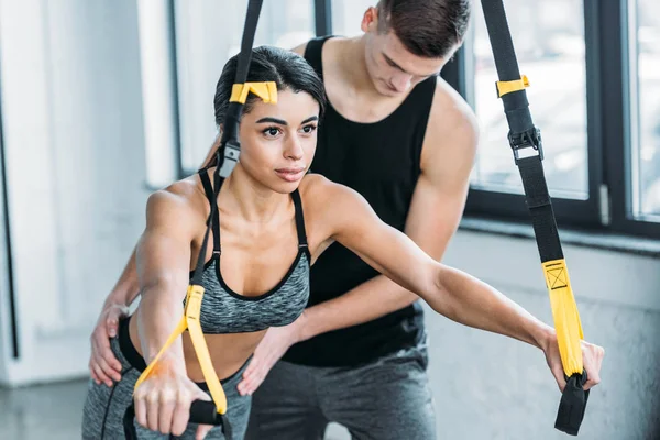 Entrenador masculino ayudando a la joven afroamericana a hacer ejercicio con bandas de resistencia en el gimnasio - foto de stock