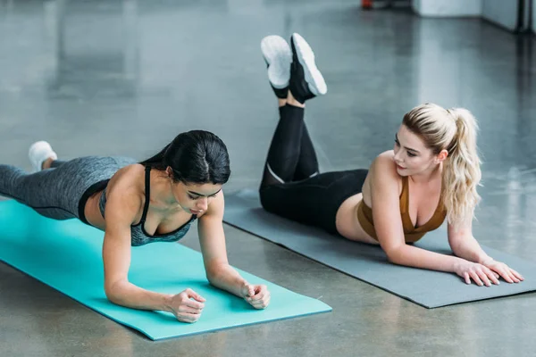 Chica rubia tumbada en la esterilla de yoga y mirando a la deportista afroamericana haciendo ejercicio de tablón en el gimnasio - foto de stock