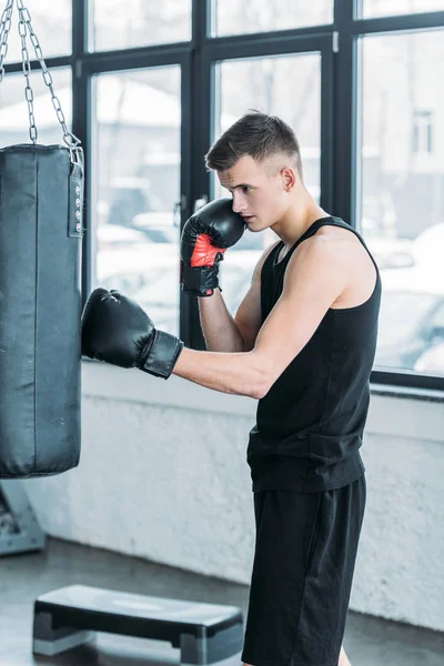 Vista lateral del joven concentrado en guantes de boxeo haciendo ejercicio con saco de boxeo en el gimnasio - foto de stock