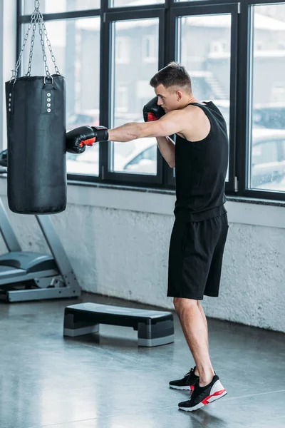 Vista lateral de jovem desportivo homem boxe com saco de perfuração no ginásio — Fotografia de Stock