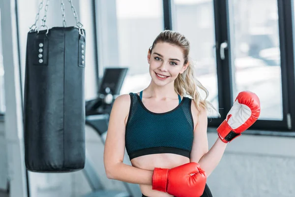 Happy beautiful sporty girl in boxing gloves smiling at camera in gym — Stock Photo