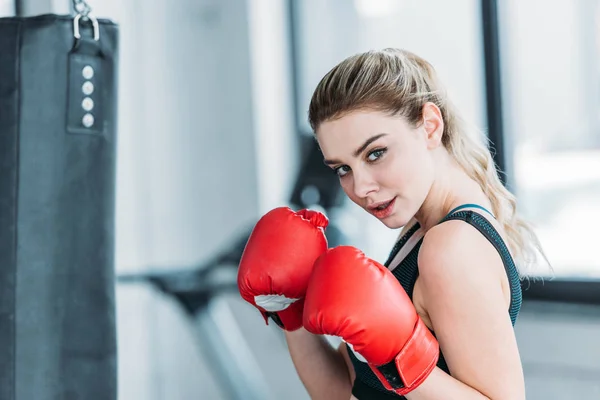 Atractiva joven deportista en guantes de boxeo mirando a la cámara en el gimnasio - foto de stock