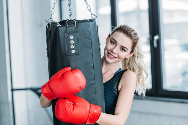 Menina desportiva feliz em luvas de boxe abraçando saco de perfuração e sorrindo para a câmera no ginásio — Fotografia de Stock