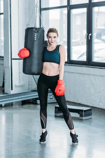 Bela menina esportiva em luvas de boxe segurando saco de perfuração e sorrindo para a câmera no ginásio — Fotografia de Stock