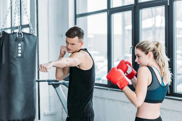 Trainer muscolare maschile e giovane donna in guanti da boxe allenamento con sacco da boxe in palestra — Foto stock