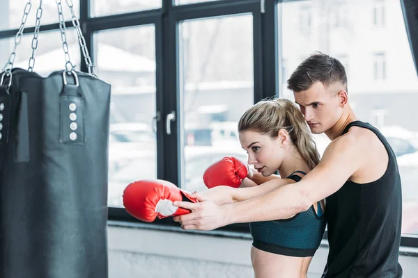 Male trainer and sporty girl in boxing gloves exercising with punching bag in gym — Stock Photo