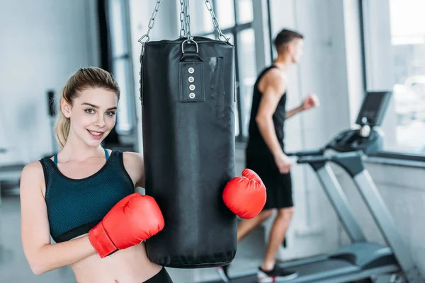 Attractive sporty girl in boxing gloves holding punching bag and smiling at camera in gym — Stock Photo