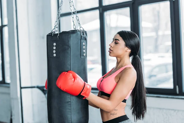 Visão lateral da jovem esportista afro-americana em luvas de boxe segurando saco de perfuração no ginásio — Fotografia de Stock