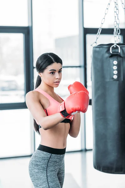 Femme afro-américaine concentrée dans l'entraînement de gants de boxe avec sac de boxe dans la salle de gym — Photo de stock