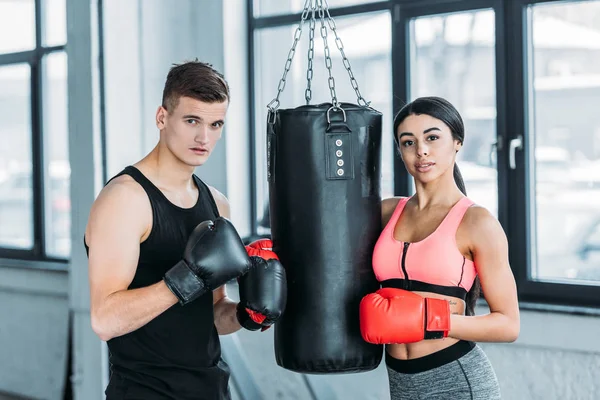Чоловічі та жіночі боксери в боксерських рукавичках, що стоять біля мішка для ударів і дивляться на камеру в спортзалі — стокове фото