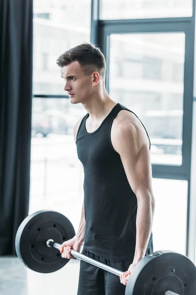 Joven deportista concentrado levantando la barra y mirando hacia otro lado en el gimnasio - foto de stock