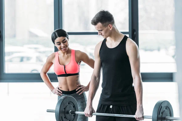 Smiling african american girl in sportswear looking at handsome young sportsman lifting barbell in gym — Stock Photo