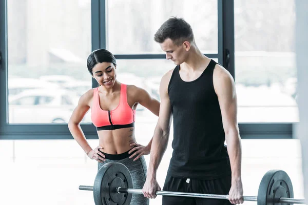 Sorridente afro-americana menina em sportswear olhando para bonito jovem desportista levantando barbell no ginásio — Fotografia de Stock