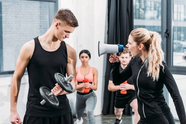 Sportswoman with megaphone yelling at young man training with dumbbell in gym — Stock Photo