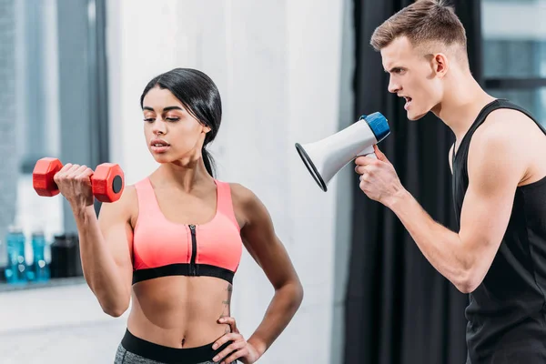 Allenatore con megafono urlando a sportivo africano americano ragazza formazione con manubri in palestra — Foto stock