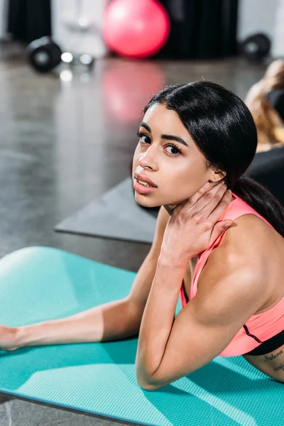 Young african american woman touching neck and looking at camera while lying on yoga mat in gym — Stock Photo