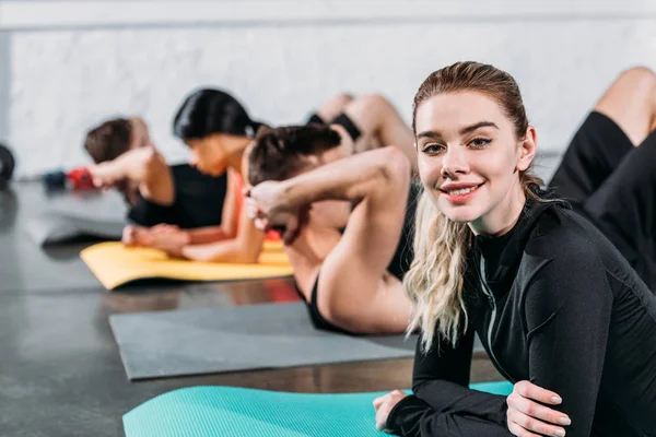 Hermosa mujer joven acostada en esterilla de yoga y sonriendo a la cámara mientras entrena con amigos en el gimnasio - foto de stock