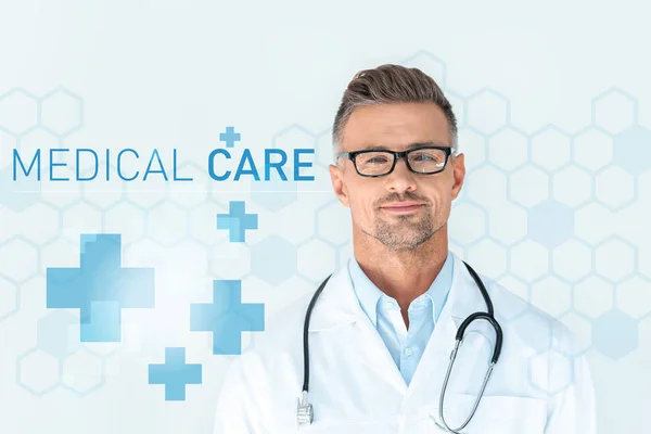 Handsome doctor in glasses with stethoscope on shoulders looking at camera with medical care lettering — Stock Photo