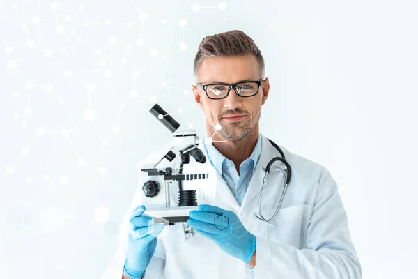 Handsome scientist in glasses holding microscope and looking at camera isolated on white with technology interface — Stock Photo