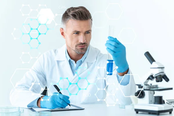 Guapo científico mirando el tubo de ensayo con reactivo azul aislado en blanco con símbolos médicos - foto de stock