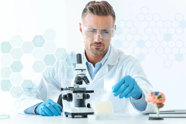 Foyer sélectif de beau scientifique dans des lunettes de protection faisant l'expérience avec le microscope isolé sur blanc avec des symboles médicaux — Photo de stock