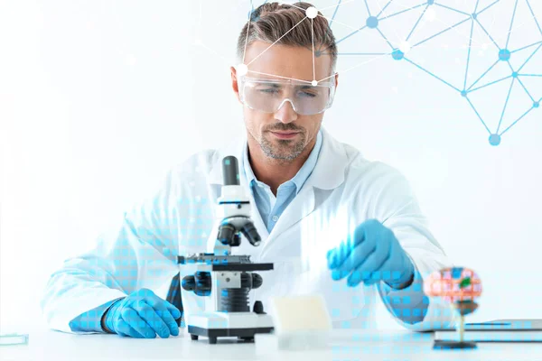 Selective focus of handsome scientist in protective glasses making experiment with microscope isolated on white with medical symbols — Stock Photo