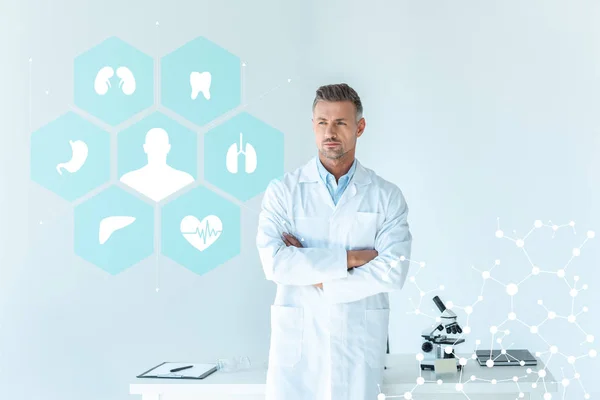 Handsome scientist standing with crossed arms near table isolated on white with medical interface — Stock Photo