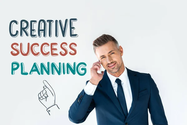 Smiling handsome businessman talking by smartphone and looking at camera isolated on white with creative success planning — Stock Photo