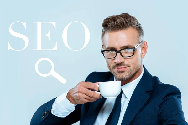Красивый бизнесмен пьет кофе и смотрит на камеру, изолированную на голубом с вывеской SEO — стоковое фото