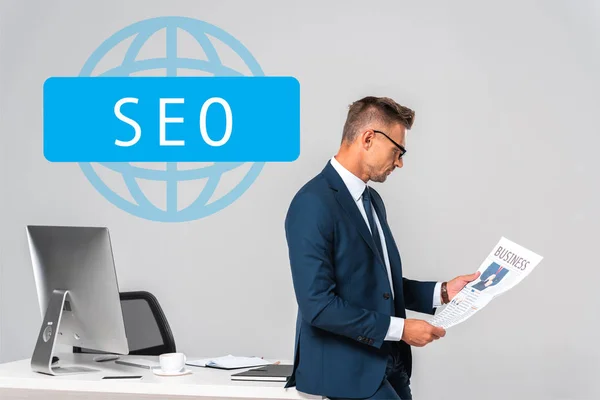 Side view of handsome businessman reading newspaper isolated on grey with seo sign — Stock Photo