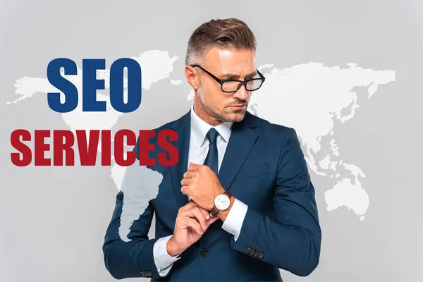 Handsome businessman wearing wristwatch and looking away isolated on grey with world map and seo services — Stock Photo