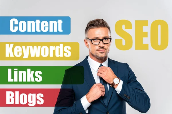 Portrait of handsome businessman in suit and glasses tying tie isolated on grey with seo lettering — Stock Photo