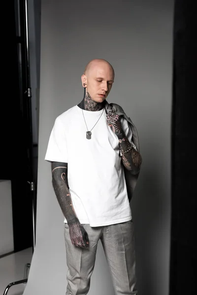 Young bald man with tattoos holding suit jacket on shoulder and standing in studio — Stock Photo