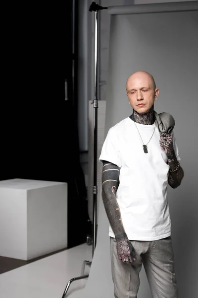 Homme chauve avec des tatouages tenant la veste de costume sur l'épaule et regardant loin en studio — Photo de stock