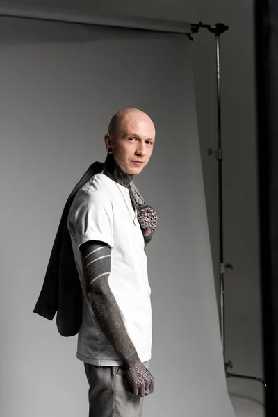 Vista lateral do homem tatuado bonito segurando jaqueta terno no ombro e sorrindo para a câmera no estúdio — Fotografia de Stock