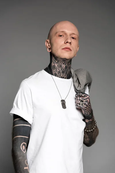 Confiado hombre tatuado en camiseta blanca sosteniendo chaqueta de traje en el hombro y mirando a la cámara aislada en gris - foto de stock