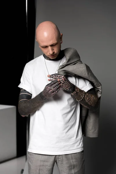 Bald tattooed man with suit jacket on shoulder looking at hands with tattoos in studio — Stock Photo