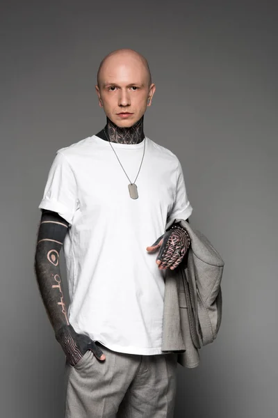 Serious bald tattooed man in white t-shirt holding suit jacket and looking at camera isolated on grey — Stock Photo