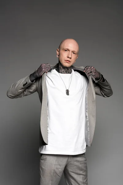 Stylish bald tattooed man wearing suit jacket and looking at camera on grey — Stock Photo