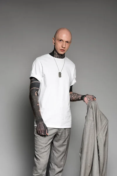 Elegante careca tatuado homem segurando casaco terno e olhando para a câmera no cinza — Fotografia de Stock