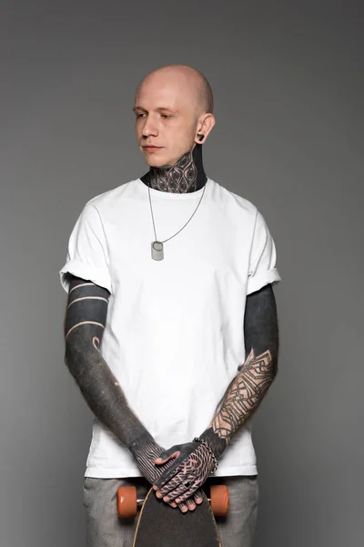 Bellissimo uomo calvo tatuato in t-shirt bianca in piedi con skateboard e distogliendo lo sguardo isolato sul grigio — Foto stock