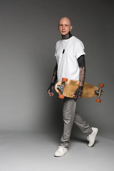 Handsome tattooed man in white t-shirt walking with longboard and looking at camera on grey — Stock Photo