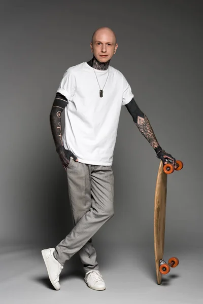 Handsome tattooed man in white t-shirt standing with skateboard and looking at camera on grey — Stock Photo