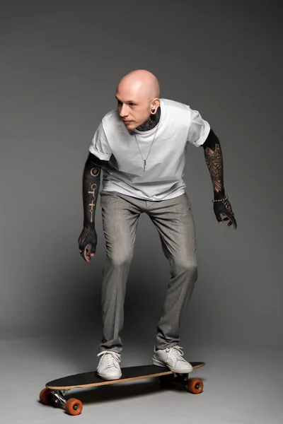 Full length view of tattooed man in white t-shirt balancing on skateboard and looking away on grey — Stock Photo