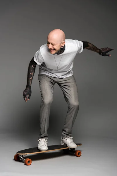 Веселый татуированный мужчина балансирует на скейтборде и смеется на сером — стоковое фото