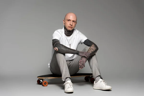 Bald tattooed man sitting on skateboard and looking at camera on grey — Stock Photo