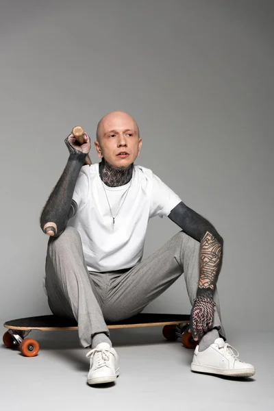 Uomo pelato tatuato seduto sullo skateboard e che tiene la mazza da baseball sul grigio — Foto stock