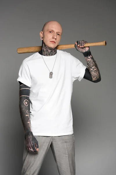 Серйозний татуйований чоловік у білій футболці тримає бейсбольну кажана і дивиться на камеру ізольовано на сірому — стокове фото