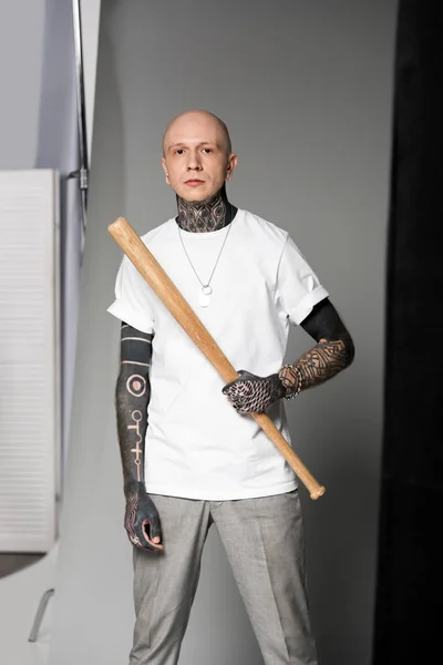 Hombre tatuado serio en camiseta blanca sosteniendo bate de béisbol y mirando a la cámara en gris - foto de stock