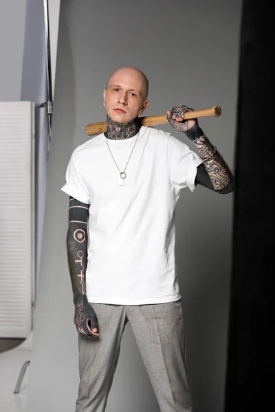 Calvo hombre tatuado en camiseta blanca de pie con bate de béisbol en el hombro y mirando a la cámara en el estudio — Stock Photo